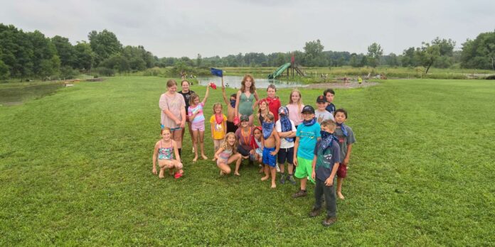 A group of smiling children and a few adults posing for a photo on a grassy field with a pond and playground equipment in the background on a cloudy day.