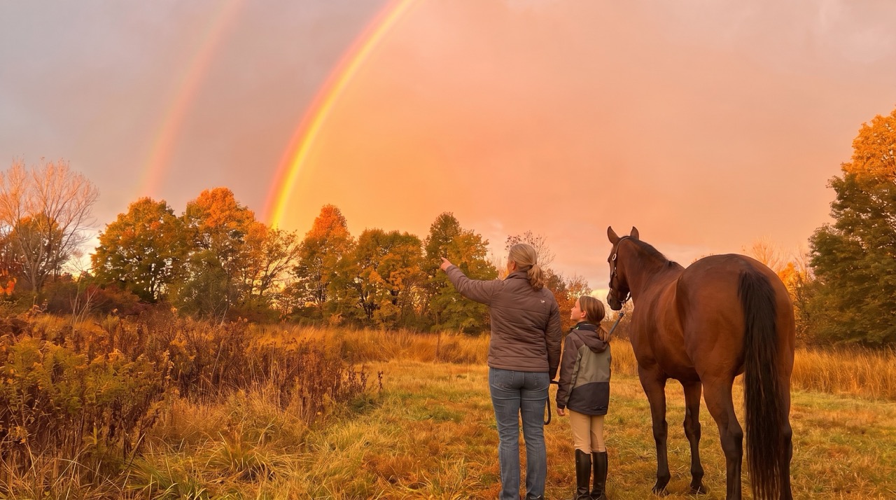 Adult and young rider standing with a horse beneath a rainbow at Autumn's Gate